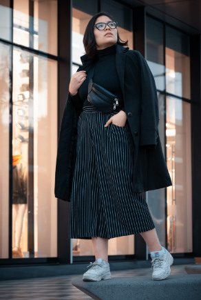 Szemüveges nő fekete culotte nadrágban és fekete kabátban egy kirakat előtt