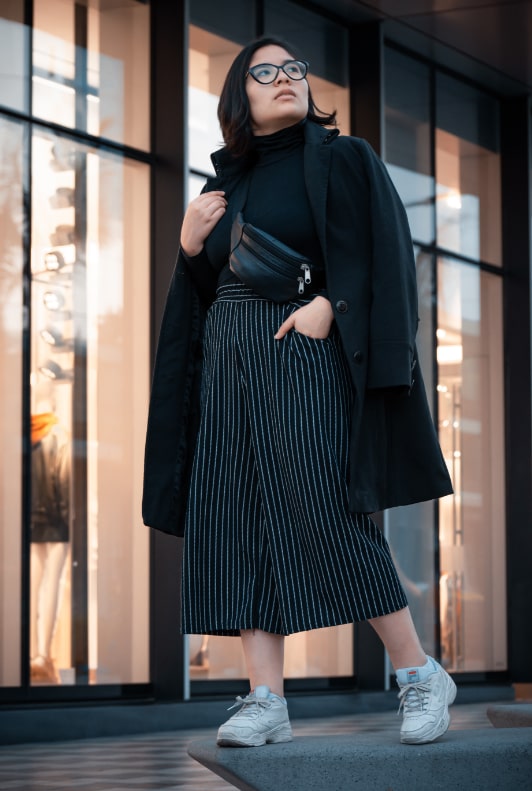 Szemüveges nő fekete culotte nadrágban és fekete kabátban egy kirakat előtt