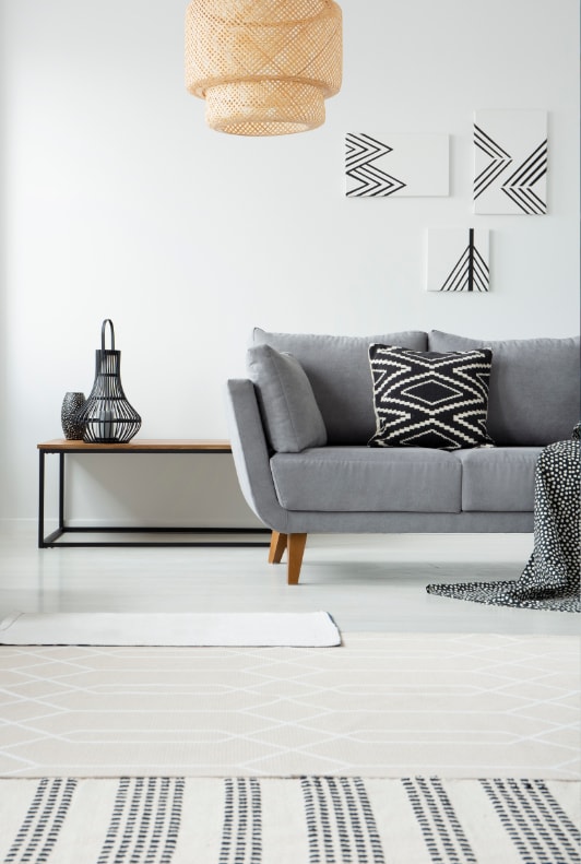 Skandináv nappaliban fehér falak, szürke kanapé, fonott lámpa, és fekete-fehér kiegészítők.