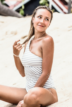 Szőke lány ül a strandon, félvállas csíkos fürdőruhában